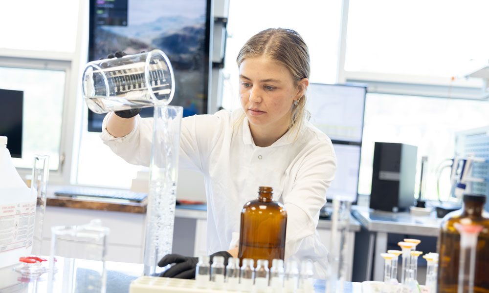 Brooklyn Becker is in a lab wearing a white lab coat. She is pouring a clear liquid from one beaker to a taller beaker to measure the volume of liquid.