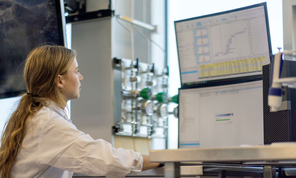 Brooklyn sits at a desk and looks at a screen full of graphs, which are produced from the contents of the test tubes.