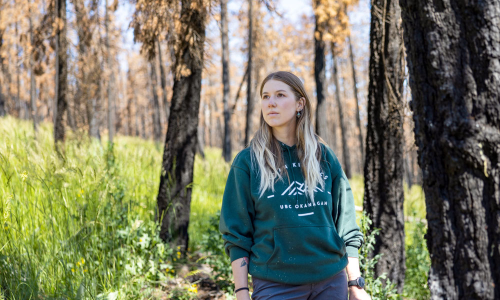 A blond female stands in the forest amidst charred trees and renewed vegetation