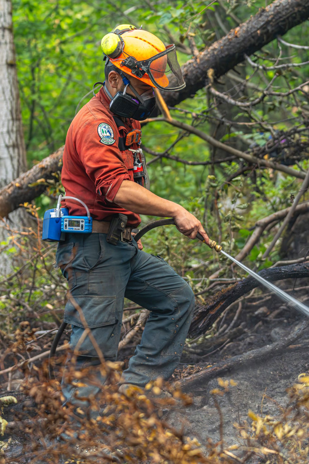 A wildland firefighter is seen in the brush wearing a respirator and spraying water on the ground.