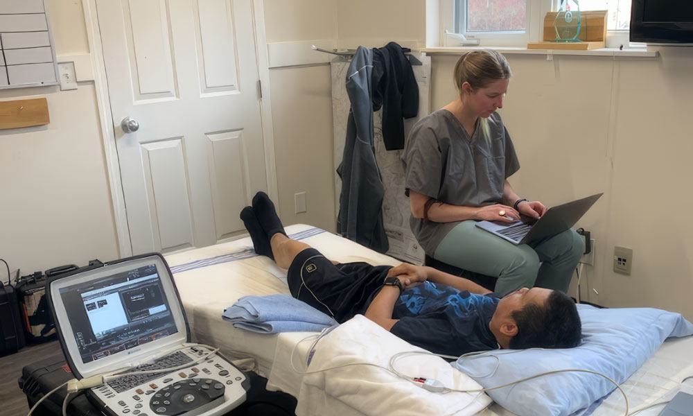 A man lies in a bed while a woman sits next to him, looking at a computer. A screen is next to the man showcasing various data.