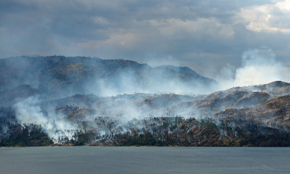 A large hillside against a lake is seen smouldering with smoke after a large forest fire.