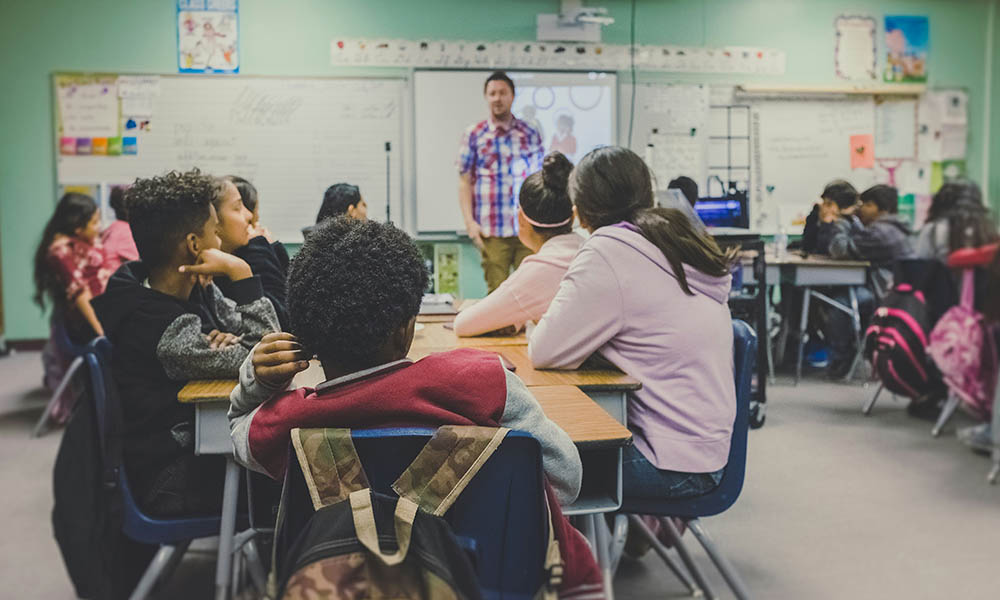 A photo of a male teacher in a plaid shirt standing in front of students sharing a study table. 