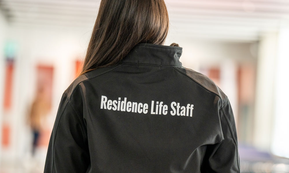 A young woman stands with her back to the camera so you can see "Residence Life Staff" written on the back of her black jacket.