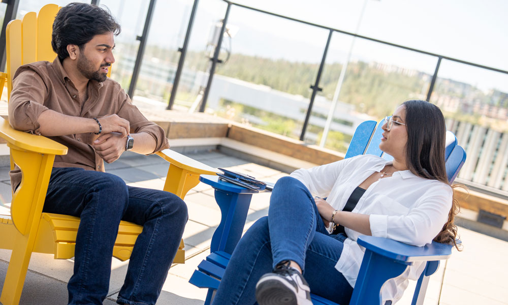 A young man and a young woman sit opposite each other on a rooftop in comfy-looking Adirondack chairs.