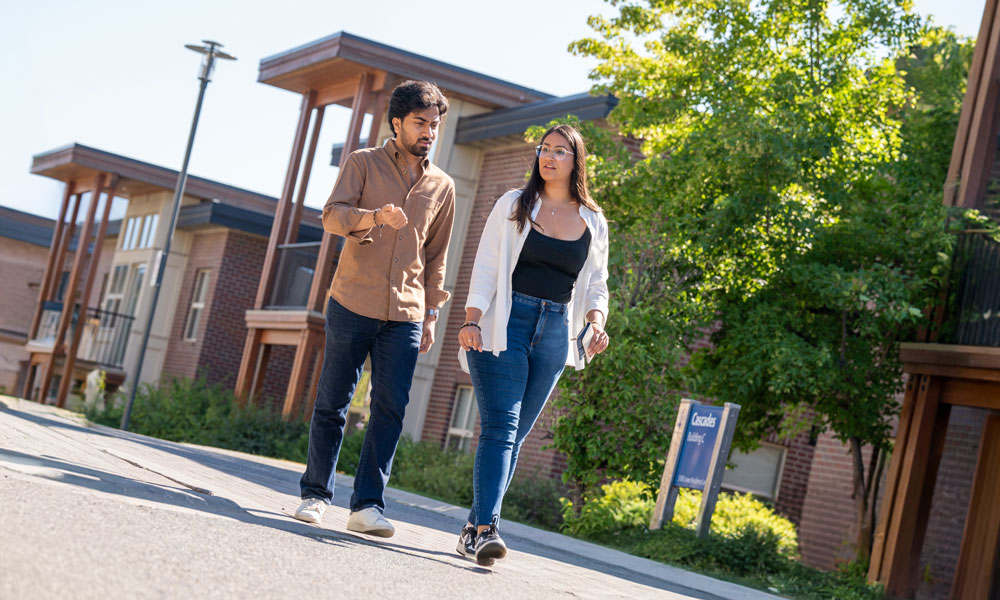 Sabhya Arora and Aradhita Arora walk side-by-side outside in the residence area of UBC Okanagan, with residence buildings in the background.