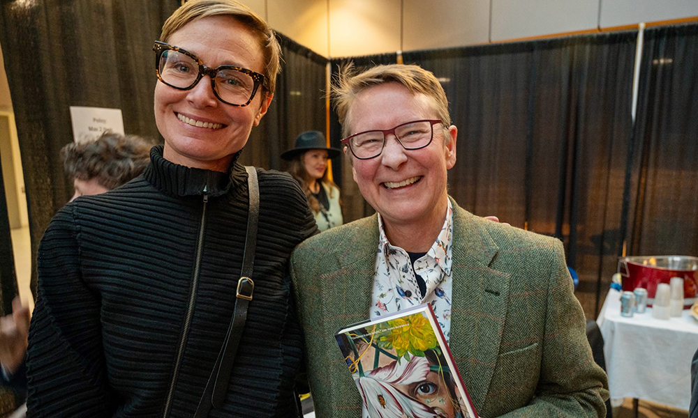 Two women smile happily at the camera while one holds a book she has written. 