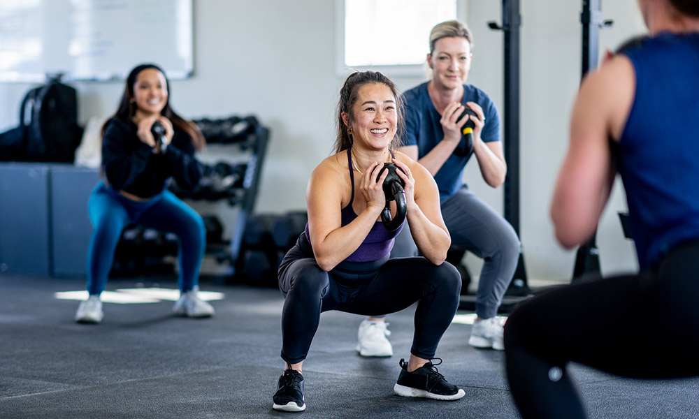 a group of people wearing workout gear are smiling while exercising with cowbell wieghts. 