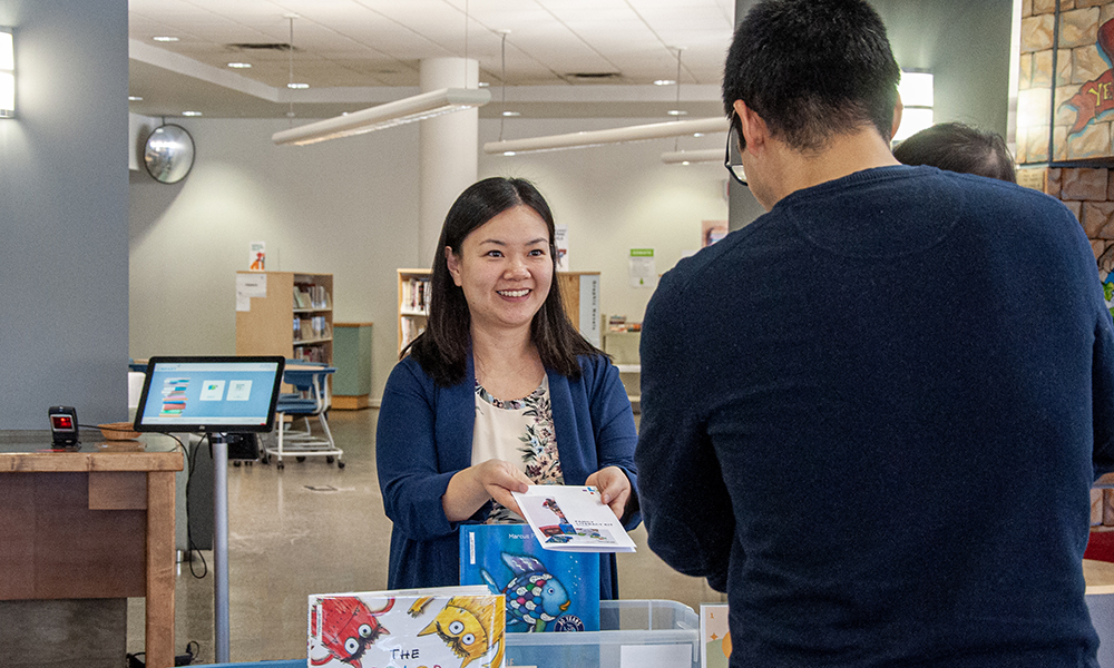 A female education professor is handing out a child literacy kit to a young father holding a baby at a public library.