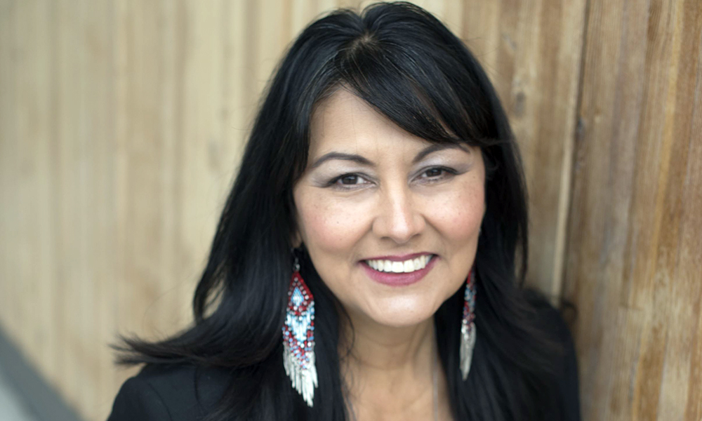 A woman with long, dark hair smiles directly into the camera. She's wearing colourful, Indigenous-themed earrings.