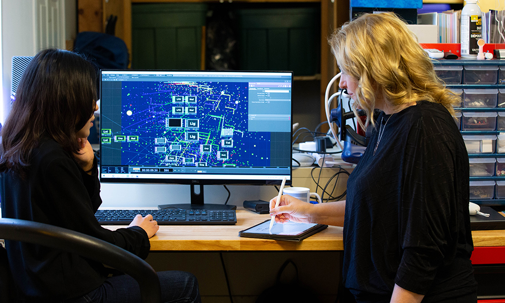 Two women sit in front of a computer screen covered with AI-generated graphics.