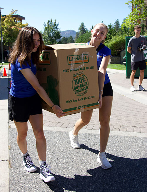 Two female members of the Heat athletic team carry a large box full of belongings