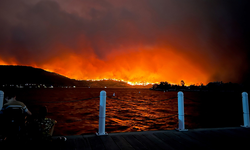 A wildfire burns out of control along the shores of Okanagan Lake during a summer evening.