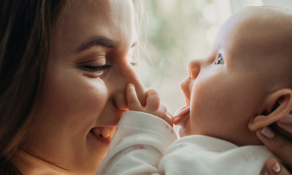 A close-up image of a mother playfully interacting with her baby. The mother smiles warmly as the baby reaches out and touches her nose with a tiny hand. The background is softly blurred, creating a tender and intimate atmosphere.