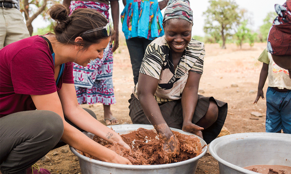 two young women have their hands in a large bowl of brown shea butter to mix it up.