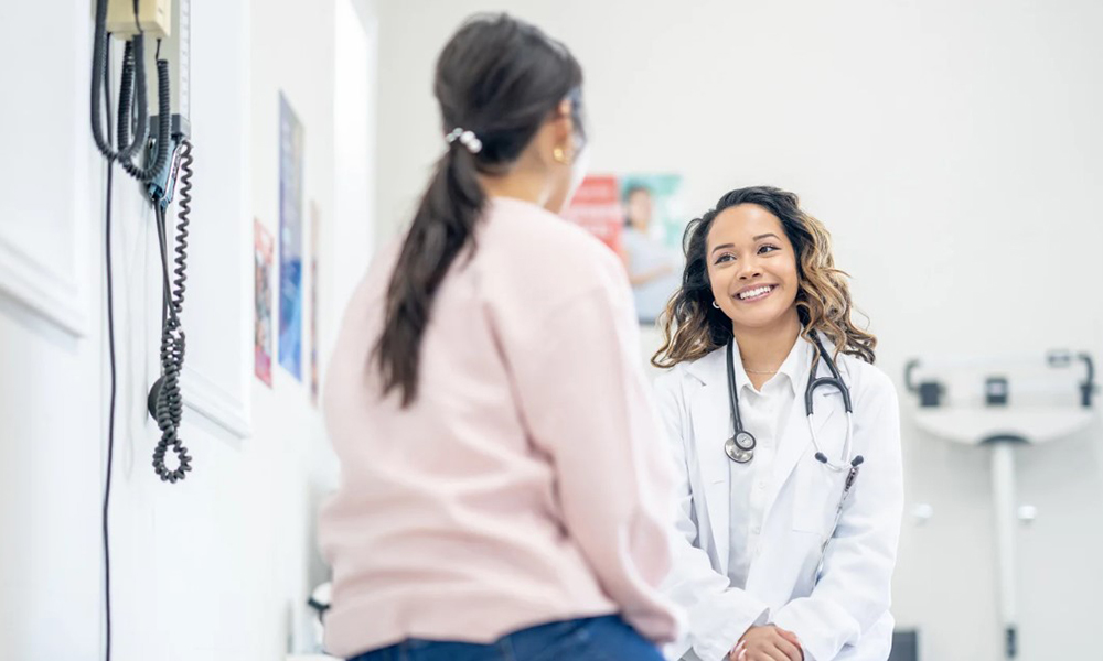 A young woman sits on an examination table and chats with a nurse. 