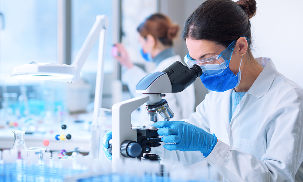 Young researcher looking at the samples under the microscope, she is wearing a face mask and protective equipment