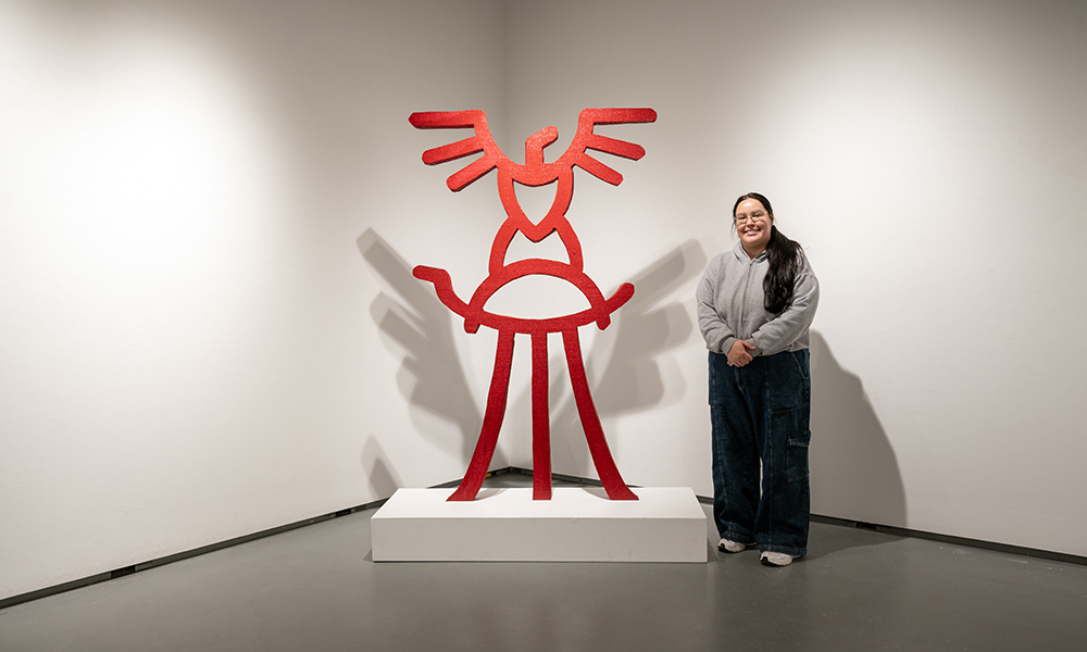 An artist stands next to a red sculpture in a starkly lit corner of an art gallery.