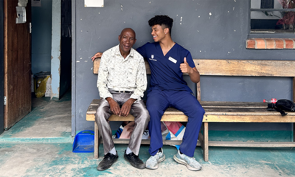 A man in scrubs sits on a bench with another man. Both smile as the man in scrubs gives a thumbs' up