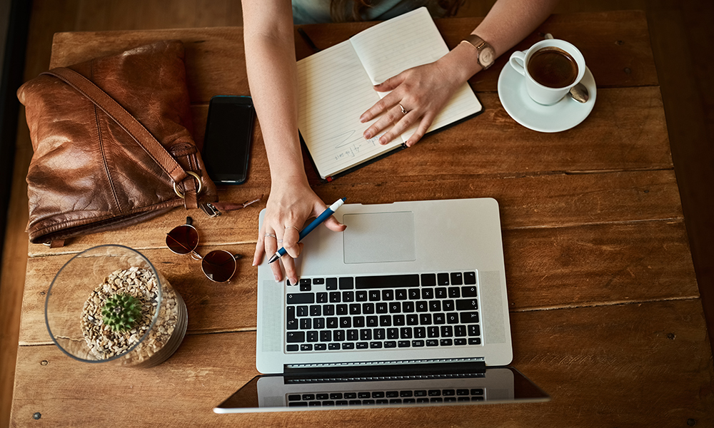 Above, hands and notebook with laptop at coffee shop as journalist for headline story or article. Person, restaurant and remote work as blogger or freelancer for creativity and research for trends.